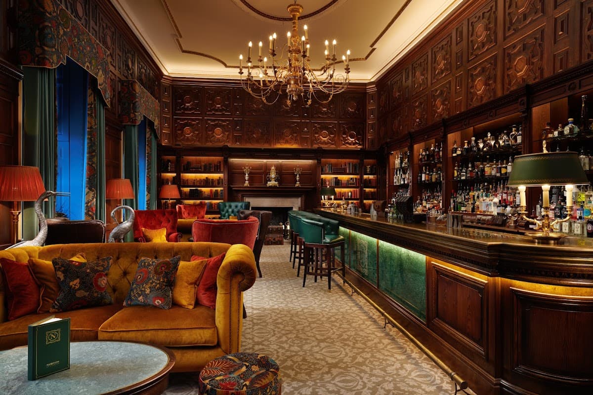 Elegant bar with ornate wood paneling, plush seating, and a well-stocked bar, illuminated by chandeliers.
