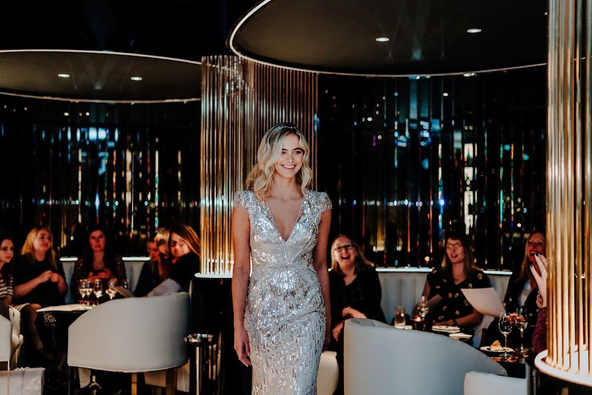 A glamorous woman in a sparkling silver dress walks through a stylish bar, with guests enjoying drinks in the background.