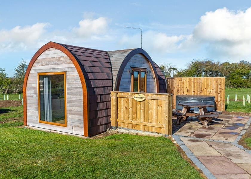 Cozy wooden holiday pod with a picnic table and fenced area, set in a green landscape under a blue sky.