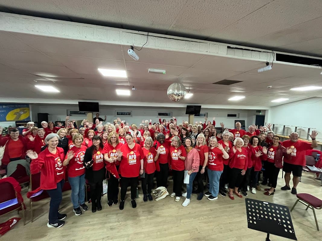 A large group of people in red shirts, smiling and waving, gathered in a spacious hall with chairs and a disco ball.