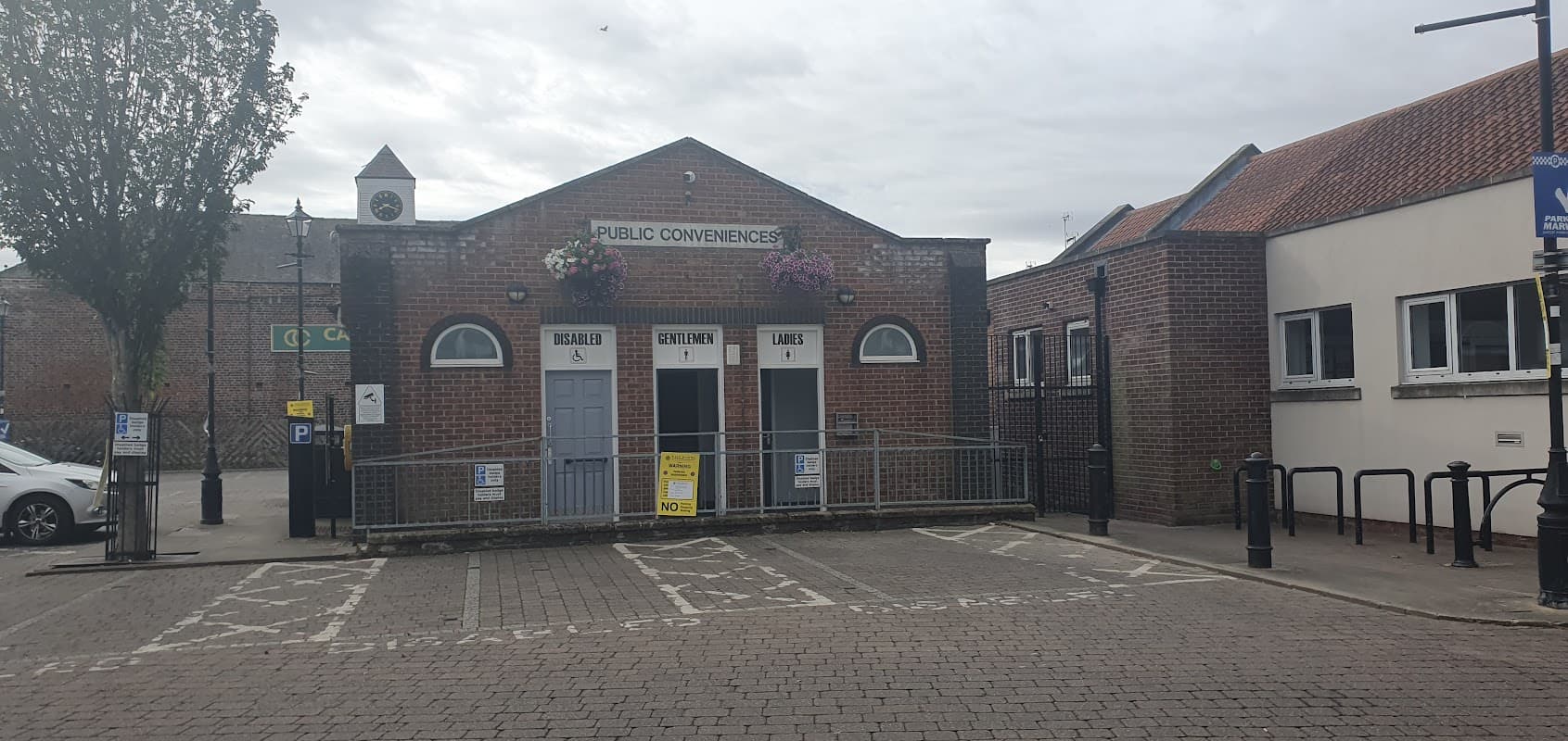 Public toilet building with three doors labeled for regulars, gentlemen, and ladies, surrounded by trees and parking.