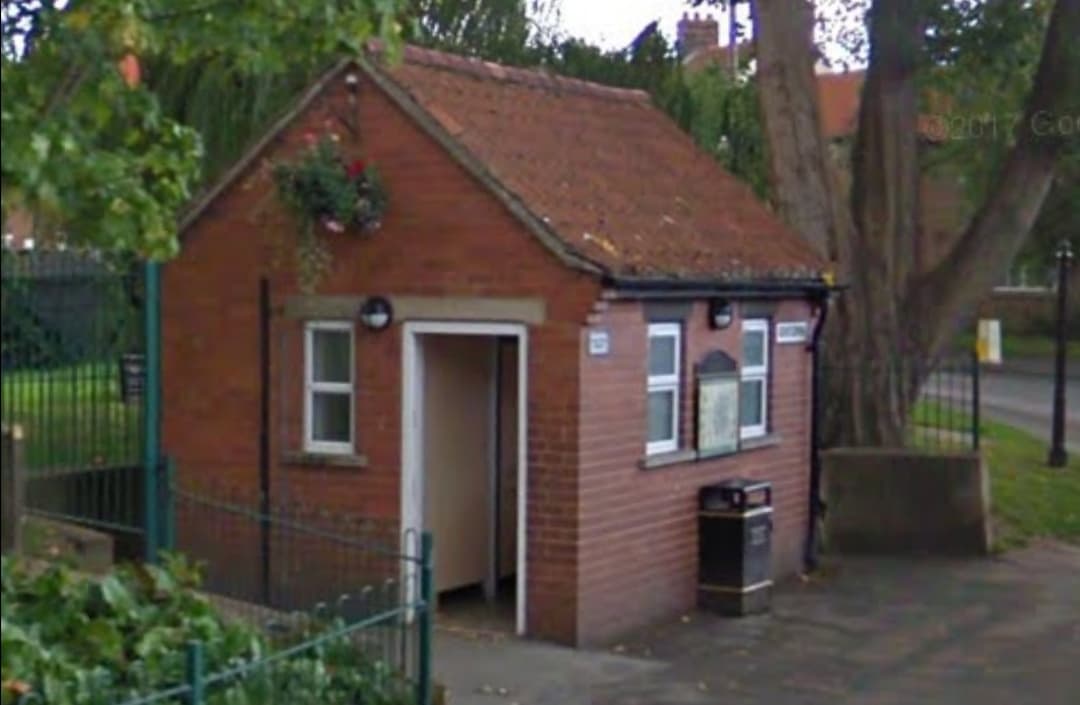 Brick building with a sloped roof, green fence, and flower pot, set in a park area.