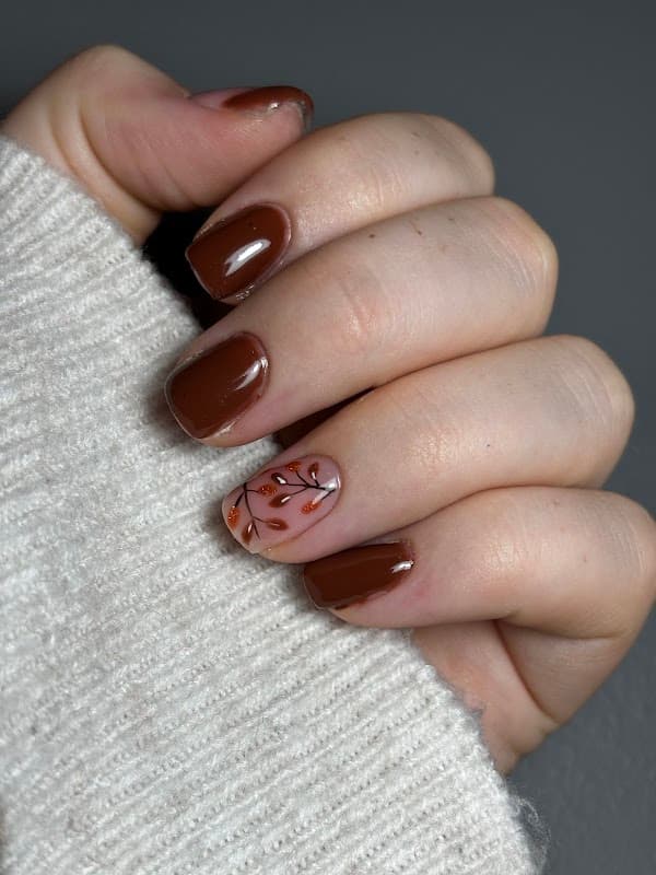 Brown manicure with a leaf design on one nail, hand resting on a light-colored sweater.
