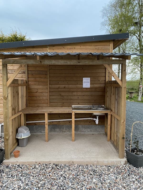 Wooden shelter with a countertop and sink, surrounded by gravel and potted plants, at Elm Lodge Holiday Park.
