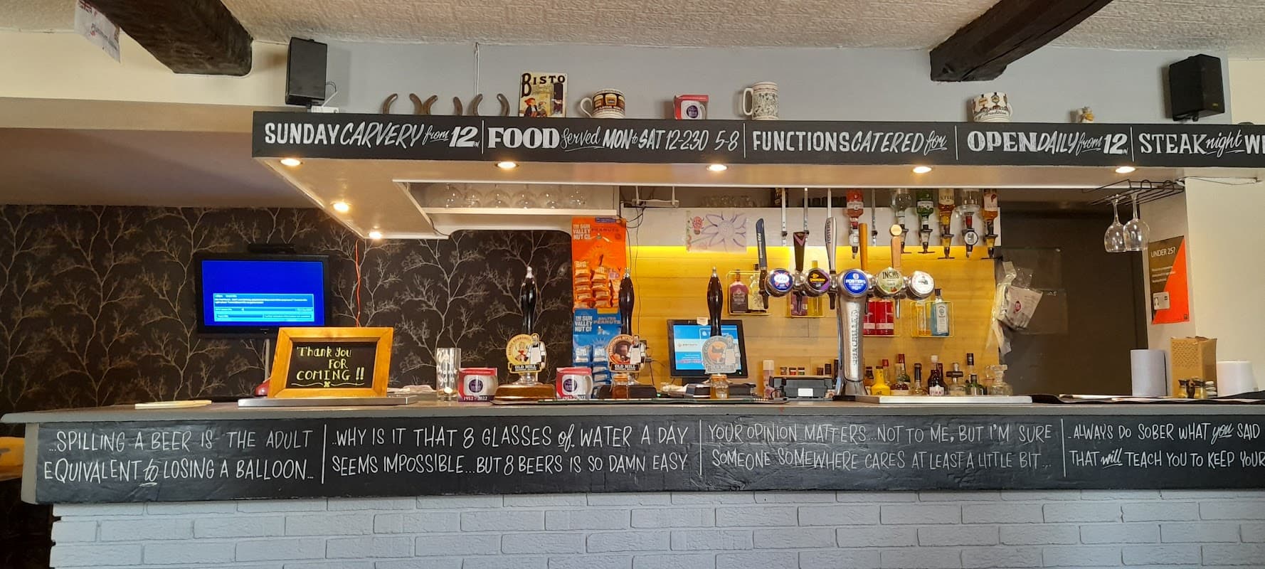 Bar counter with taps, beer glasses, and a menu board; cozy interior with wooden beams and decorative wallpaper.