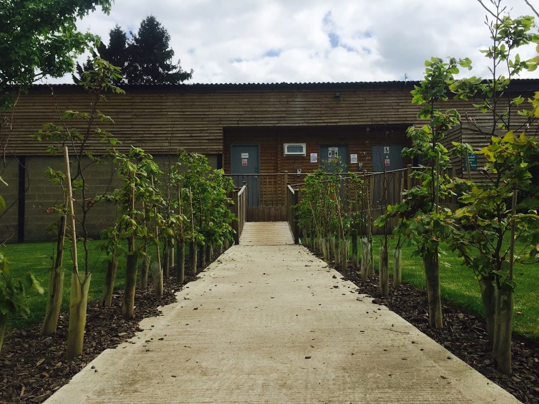 Pathway lined with young trees leading to a wooden building at The Burrows Country Park, surrounded by green grass.