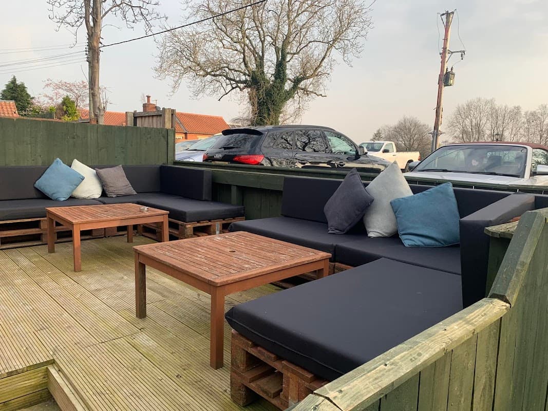 Outdoor seating area with dark sofas and wooden tables, surrounded by trees and parked cars in Great Smeaton, Yorkshire.
