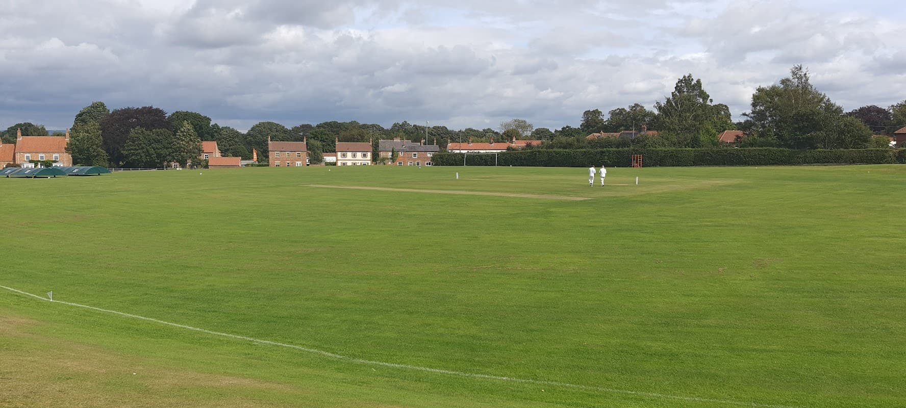 Village hall in Green Hammerton with cricket players on a lush green field under a cloudy sky.