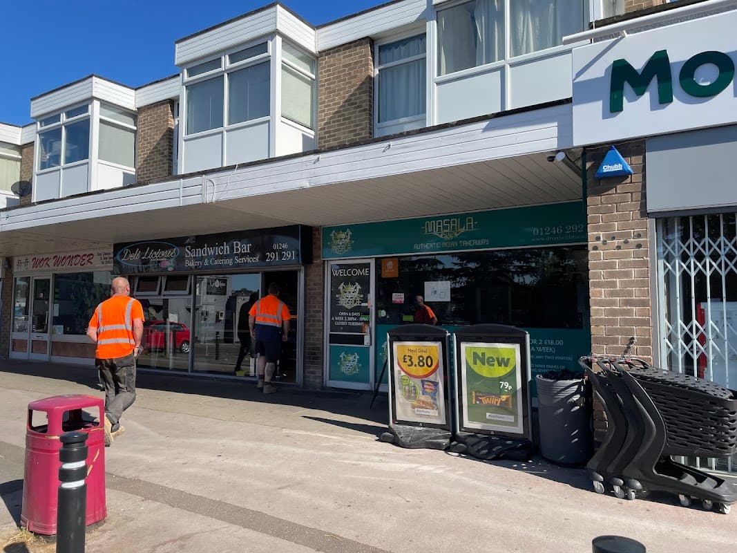 Brightly lit car park with shops, including a sandwich bar, and two workers in high-visibility vests.
