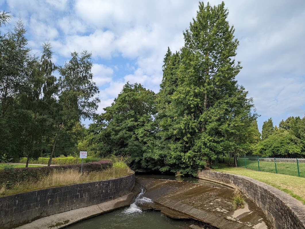Lush greenery surrounds a narrow waterway, with a stone bank and a sign visible nearby under a partly cloudy sky.
