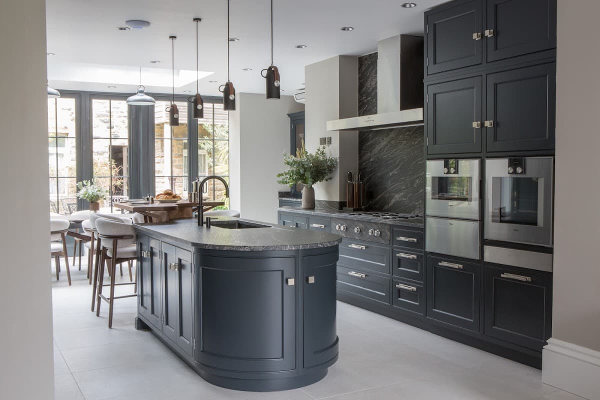 Modern kitchen with dark cabinetry, marble accents, and an island, featuring pendant lights and large windows.