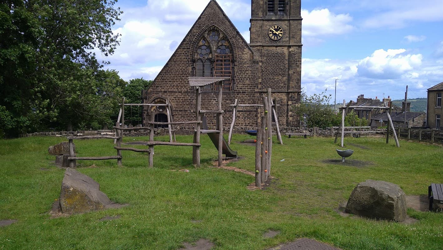 Playground equipment in a grassy area near a historic stone church with a clock tower and cloudy blue sky.