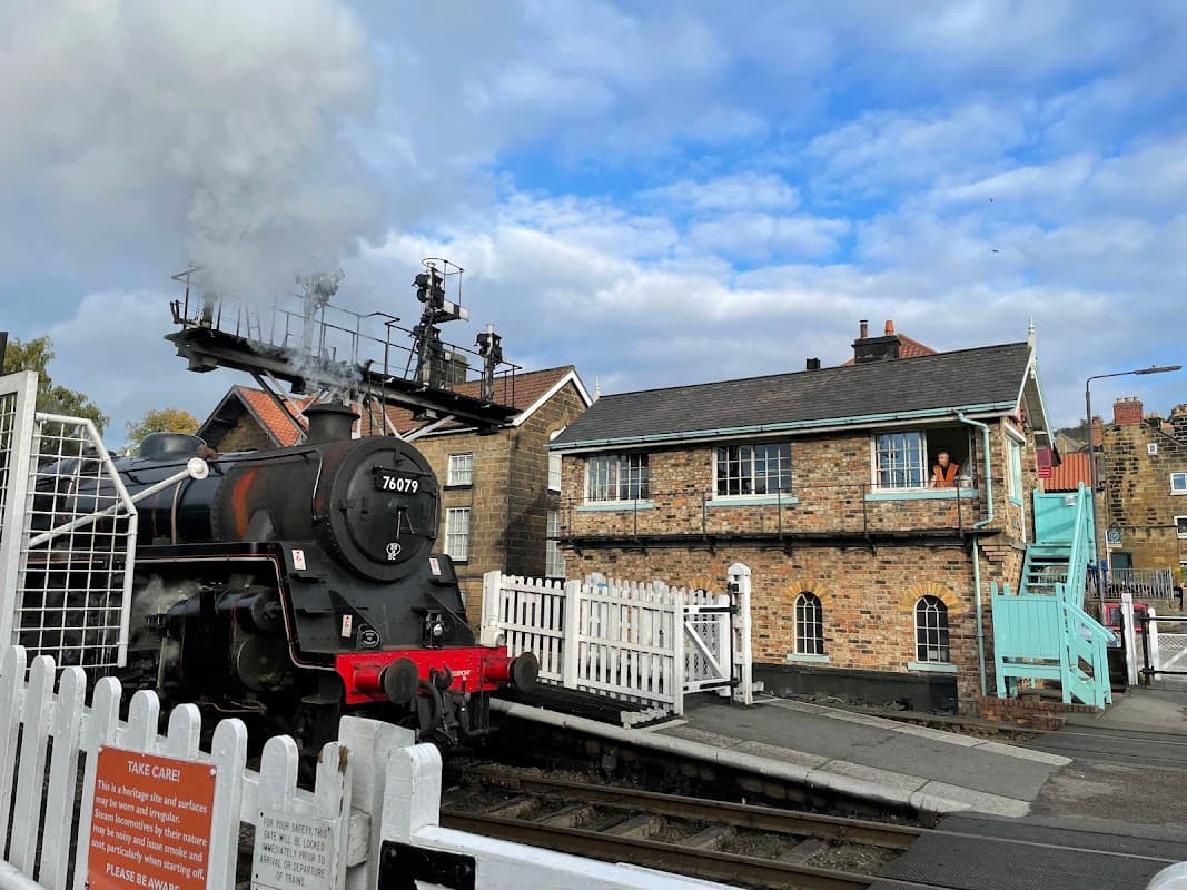 Steam train passing through Grosmont Car & Coach Park, with a historic building and cloudy sky in the background.