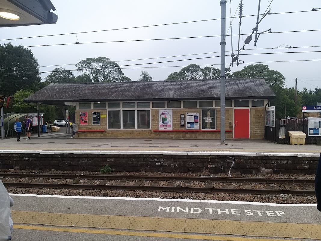 Guiseley Station with a stone building, posters, and a "Mind the Step" warning on the platform. Trees in the background.