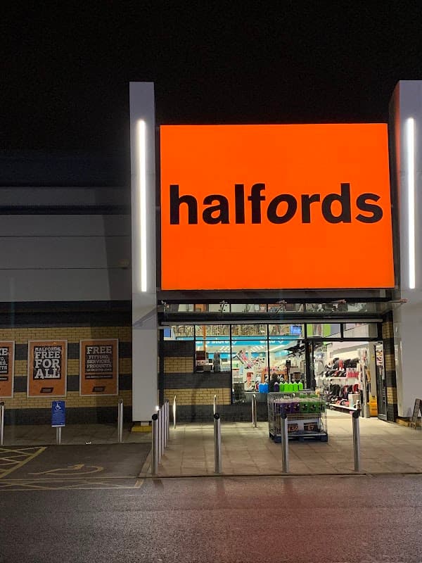 Brightly lit Halfords store entrance at night, featuring large orange signage and a display of various products outside.