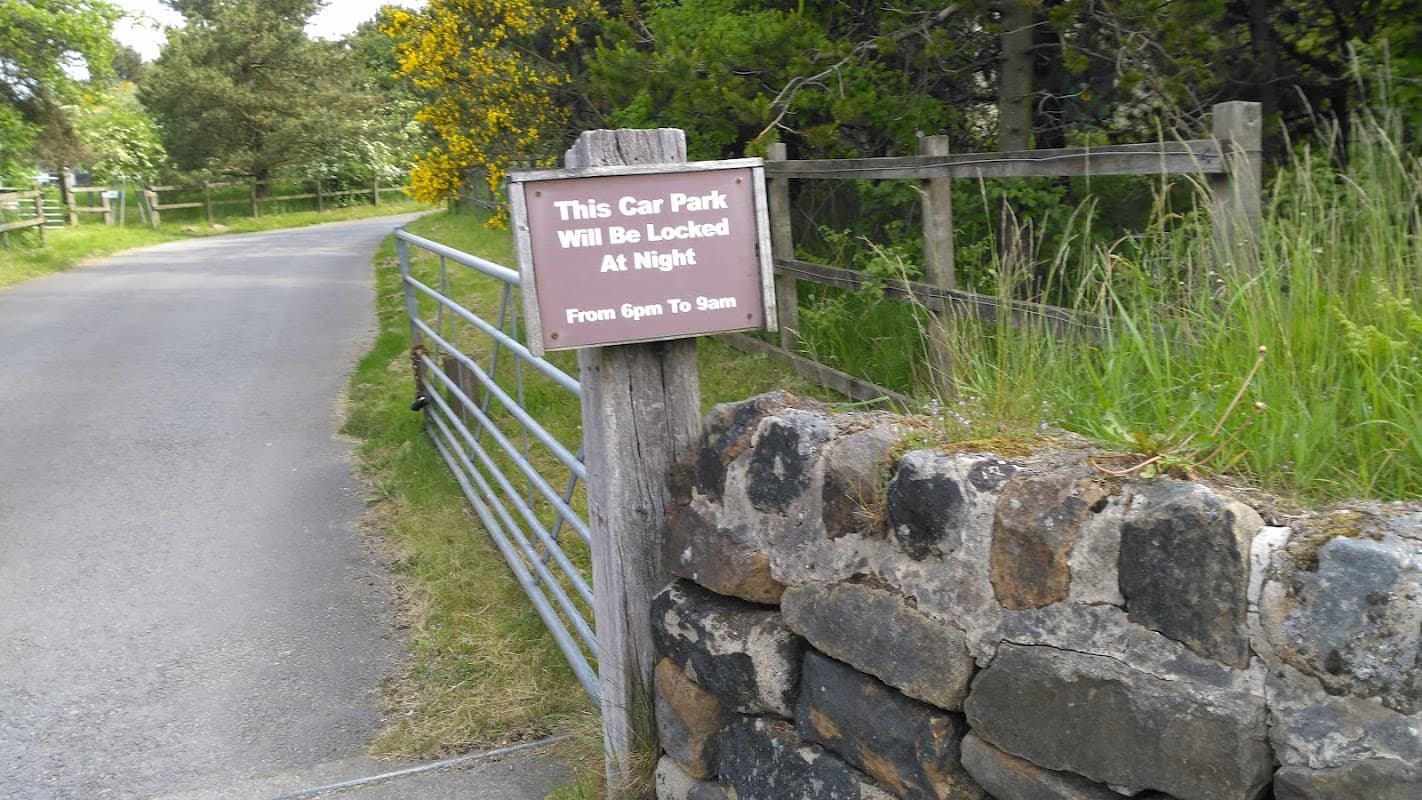 Sign indicating car park hours, surrounded by greenery and a stone wall, with a gravel road leading in.
