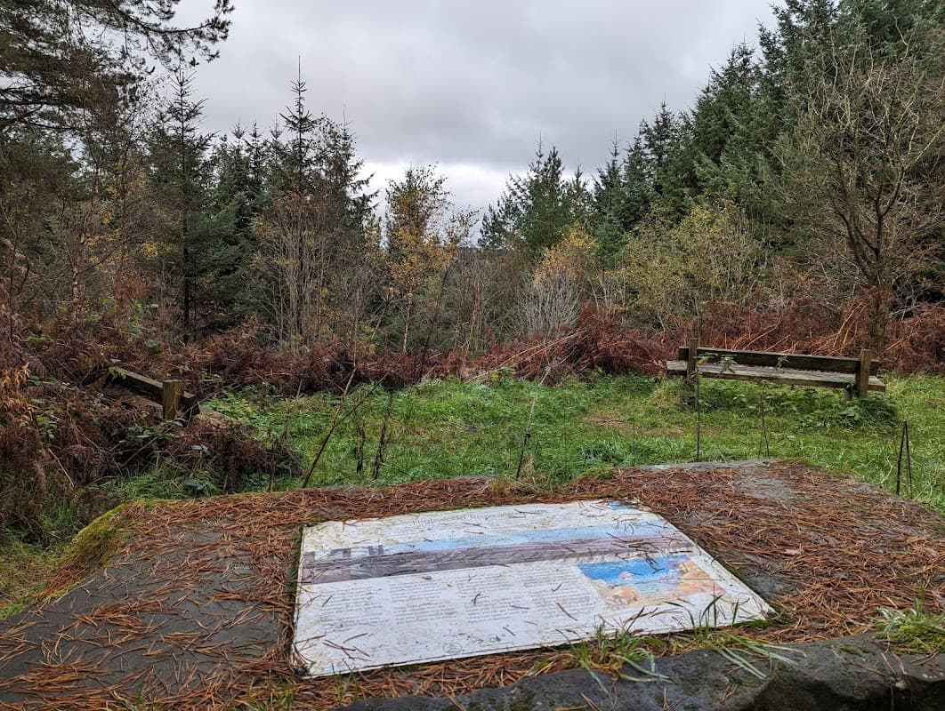 Sign on a stone slab with a map, surrounded by trees and overgrown vegetation, with benches in the background.