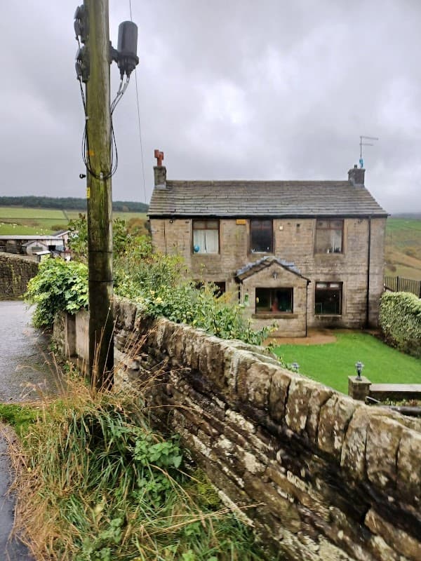 Stone building with a slate roof, surrounded by greenery and a dry stone wall, under a cloudy sky.