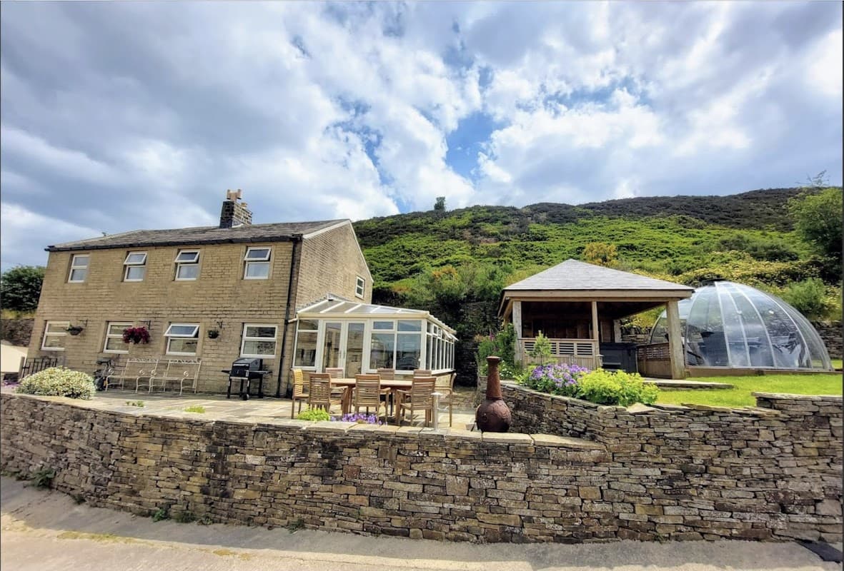Stone farmhouse with a glass conservatory, outdoor seating, and a dome structure, set against a hilly backdrop.
