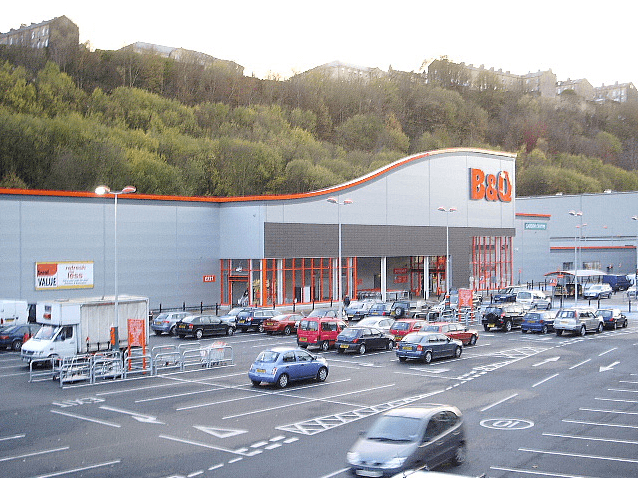 B&Q store exterior with large signage, surrounded by a parking lot filled with cars and wooded hills in the background.