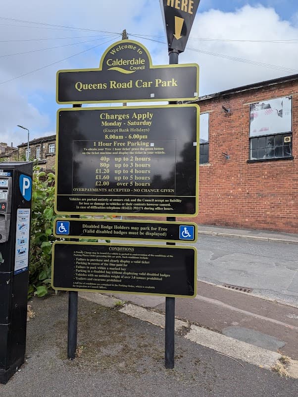 Sign for Queens Road car park with parking charges and conditions, surrounded by buildings and a road in Halifax, Yorkshire.