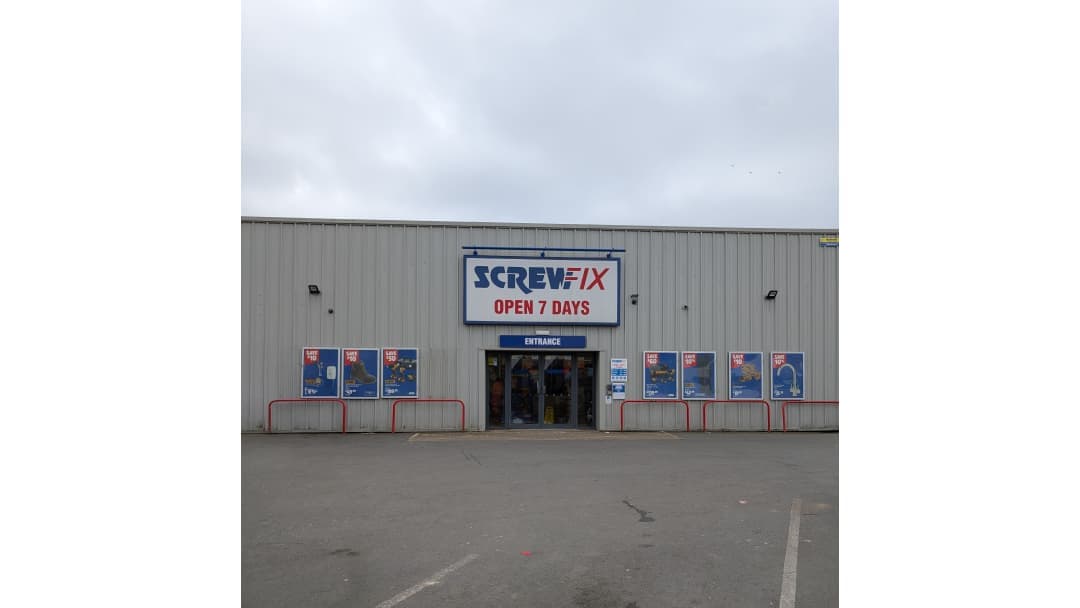 Screwfix store front with large sign, entrance doors, and promotional posters on windows, set against a cloudy sky.
