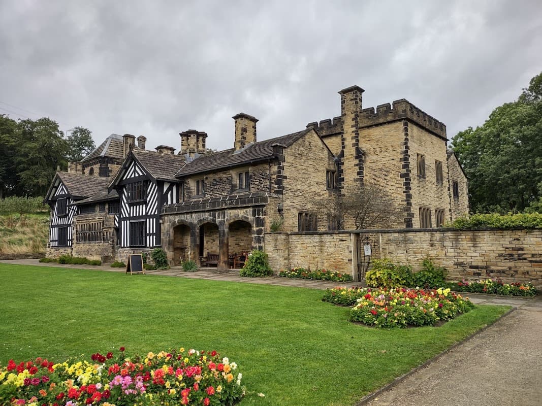 Historic Shibden Hall with a mix of stone and timber architecture, surrounded by a lush green lawn and colorful flower beds.