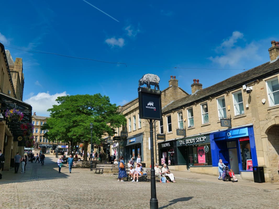 Woolshops shopping area in Halifax, featuring stone buildings, a vibrant tree, and shoppers enjoying a sunny day.