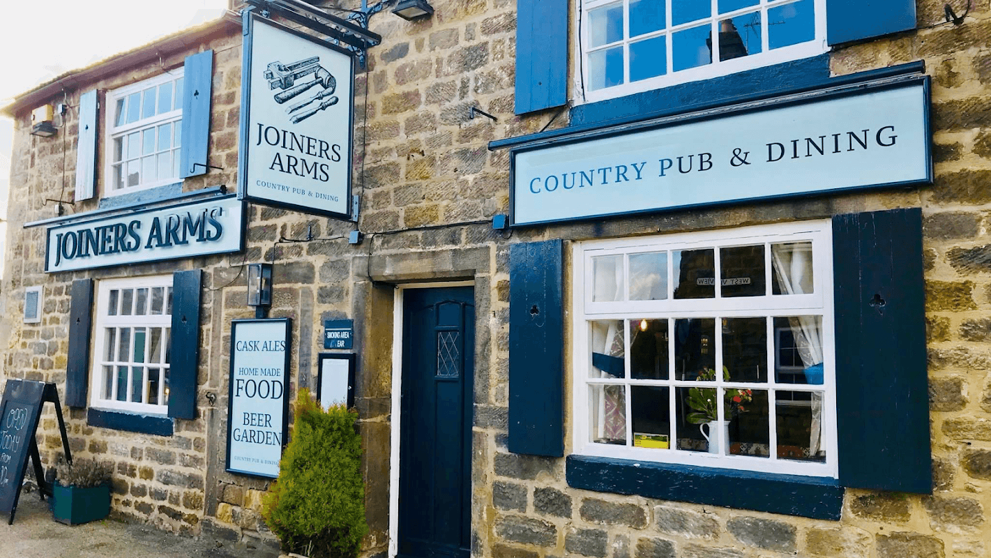 Historic stone pub with blue shutters, signs for "Joiners Arms" and "Country Pub & Dining," and a welcoming entrance.