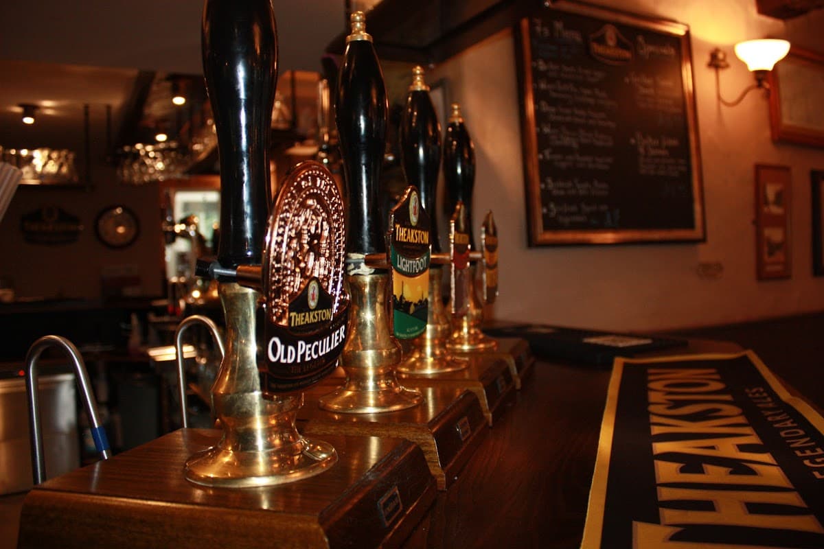 Four beer taps with distinctive labels, a wooden bar surface, and a chalkboard menu in a cozy pub setting.
