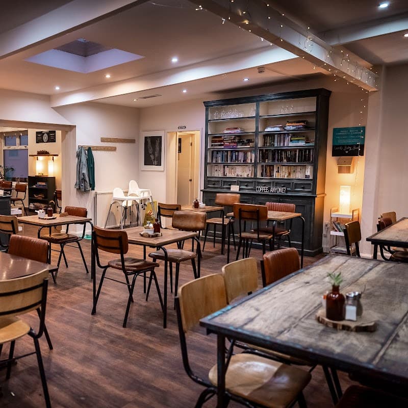 Cozy cafe interior with wooden tables, chairs, and a bookshelf, illuminated by soft lighting and a skylight.