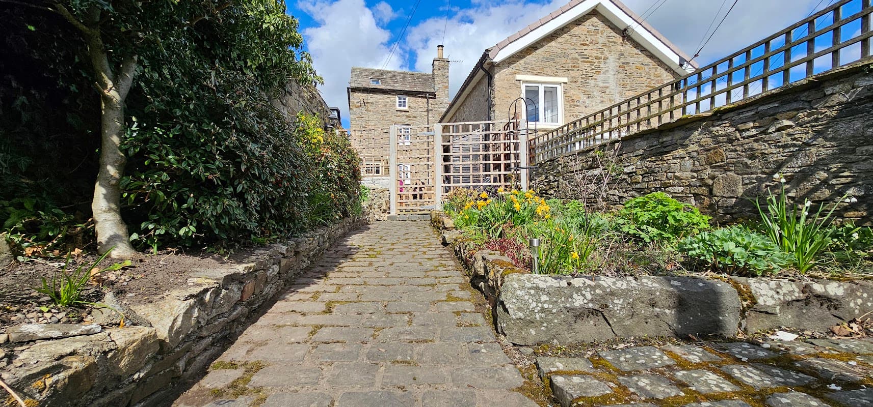 Pathway leading to Puzzle Cottage, flanked by stone walls and vibrant garden flowers under a blue sky.