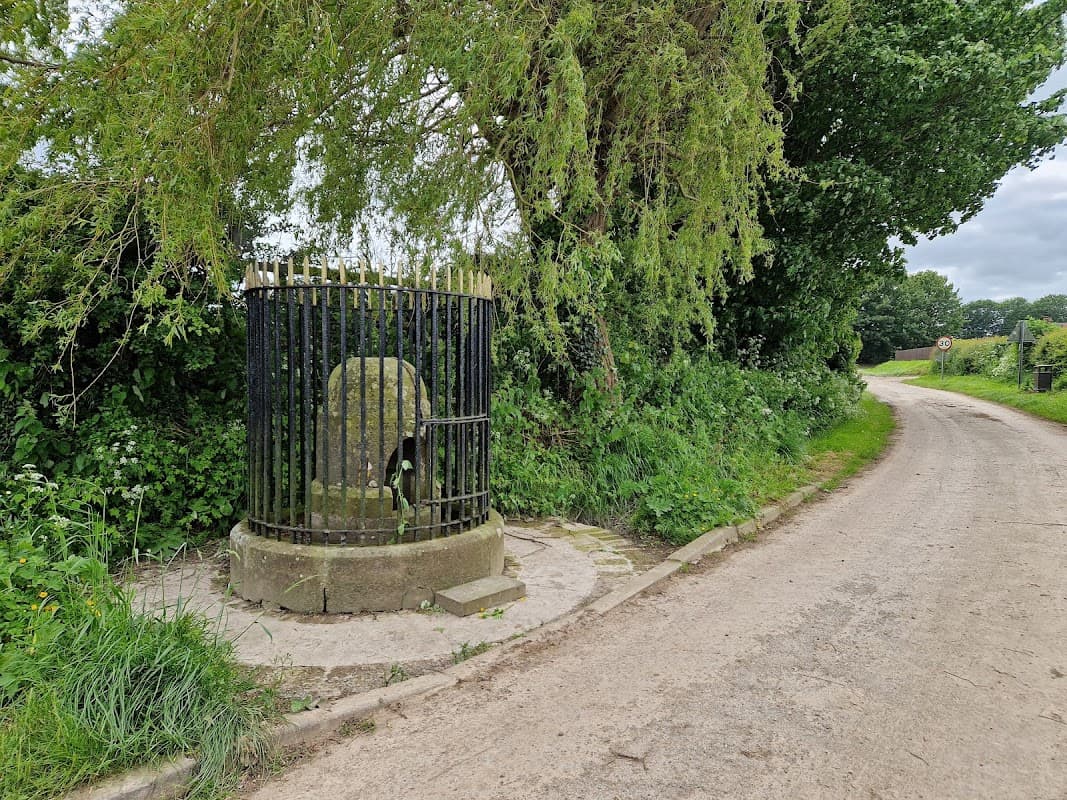 Historic stone well with a black metal railing, surrounded by greenery and a dirt road in Harpham, Yorkshire.