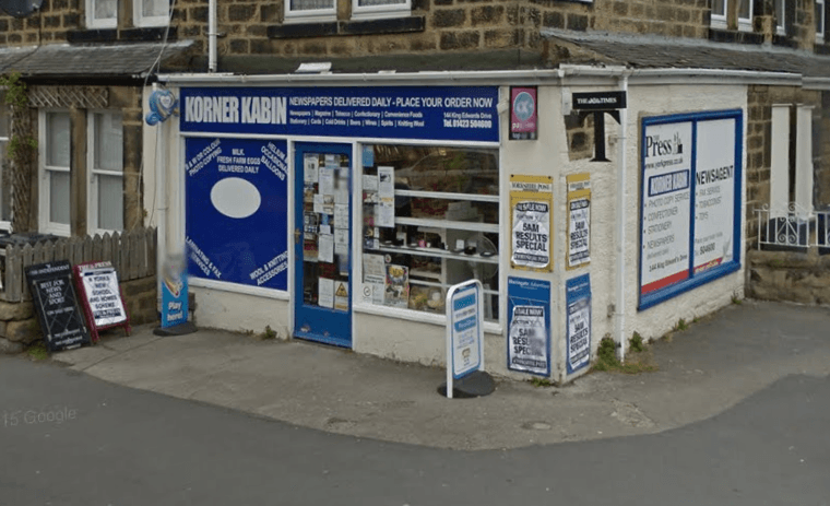 Local off-licence with a blue sign, window displays, and a sandwich board outside, set in a stone building.