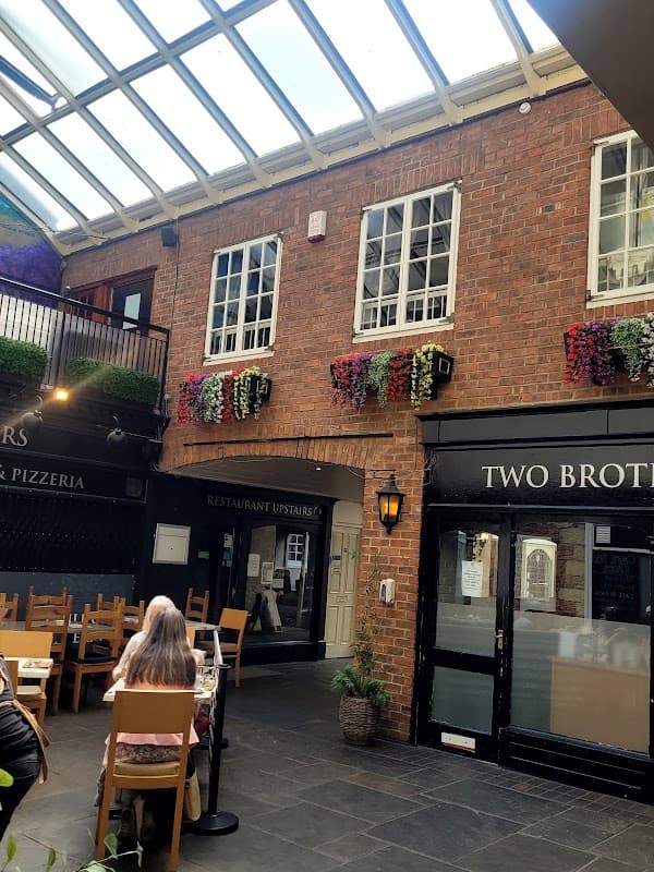 Brick walls adorned with flower baskets, glass ceiling, wooden chairs, and restaurant entrances in a cozy courtyard setting.