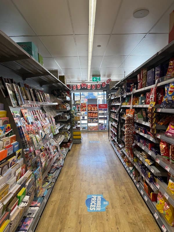 Aisle in a Co-op Food store with shelves stocked with magazines, snacks, and drinks, leading to the entrance.