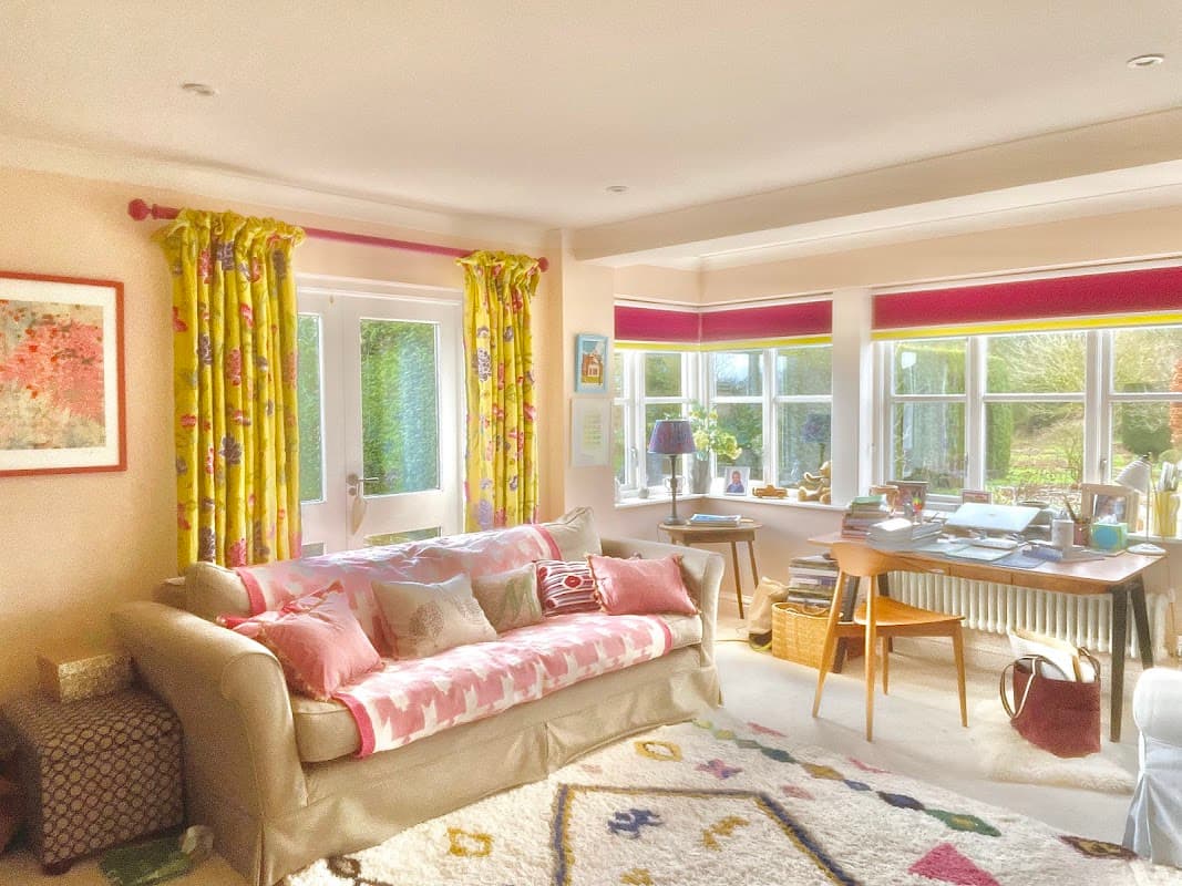 Bright living room with floral curtains, a cozy sofa, a colorful rug, and a desk by large windows.