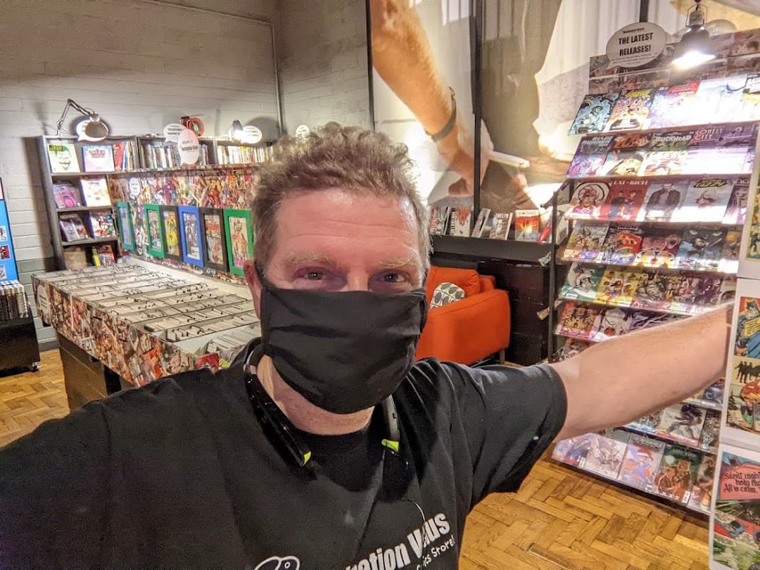 A man in a black mask stands in a vibrant bookshop filled with comics, colorful displays, and a cozy orange sofa.