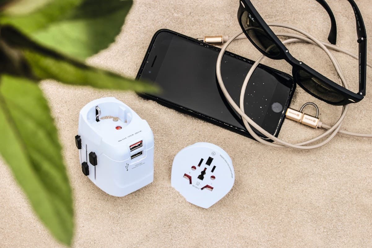 White travel adapter, smartphone, earphones, and sunglasses on sandy surface with green leaves in the corner.