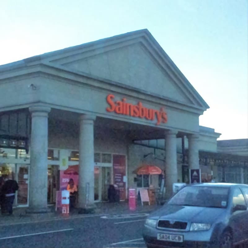 Sainsbury's storefront with columns, featuring Argos signage, shopping carts, and a parked car outside.