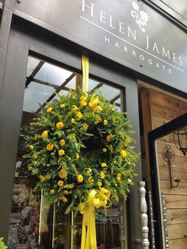 A vibrant yellow floral wreath with a bow, hanging on a shop door under a sign that reads "Helen James Harrogate."