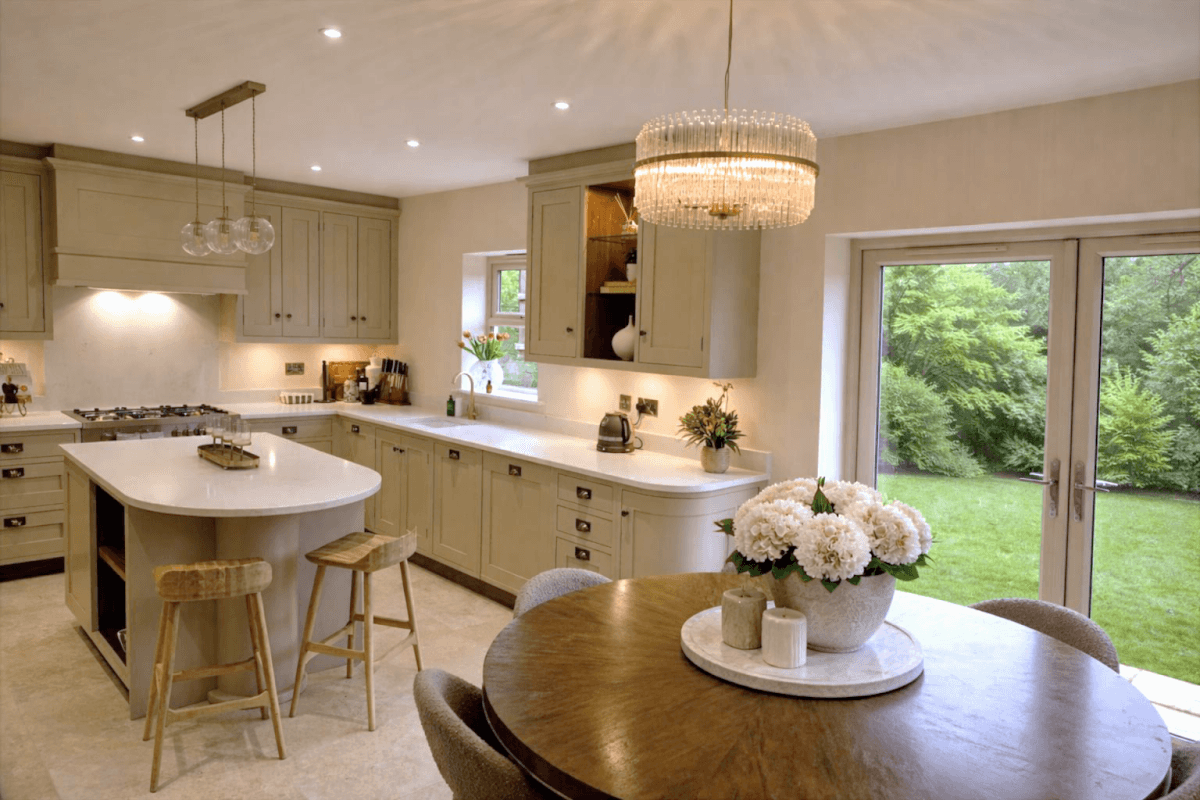Modern kitchen with light cabinetry, an island, and a round dining table adorned with flowers, overlooking a garden.