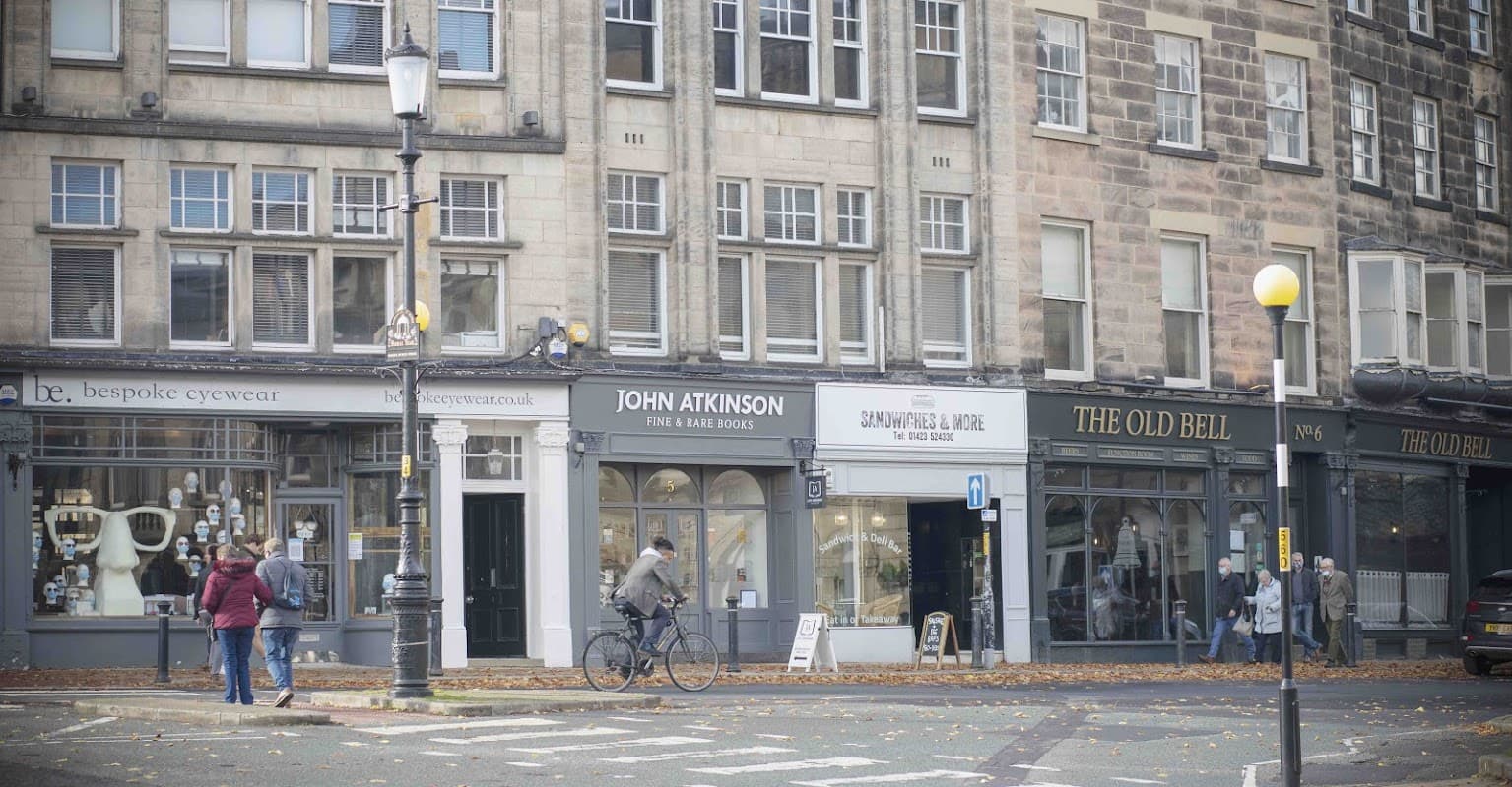 John Atkinson Fine & Rare Books storefront, with pedestrians and a cyclist on a tree-lined street in Harrogate, Yorkshire.
