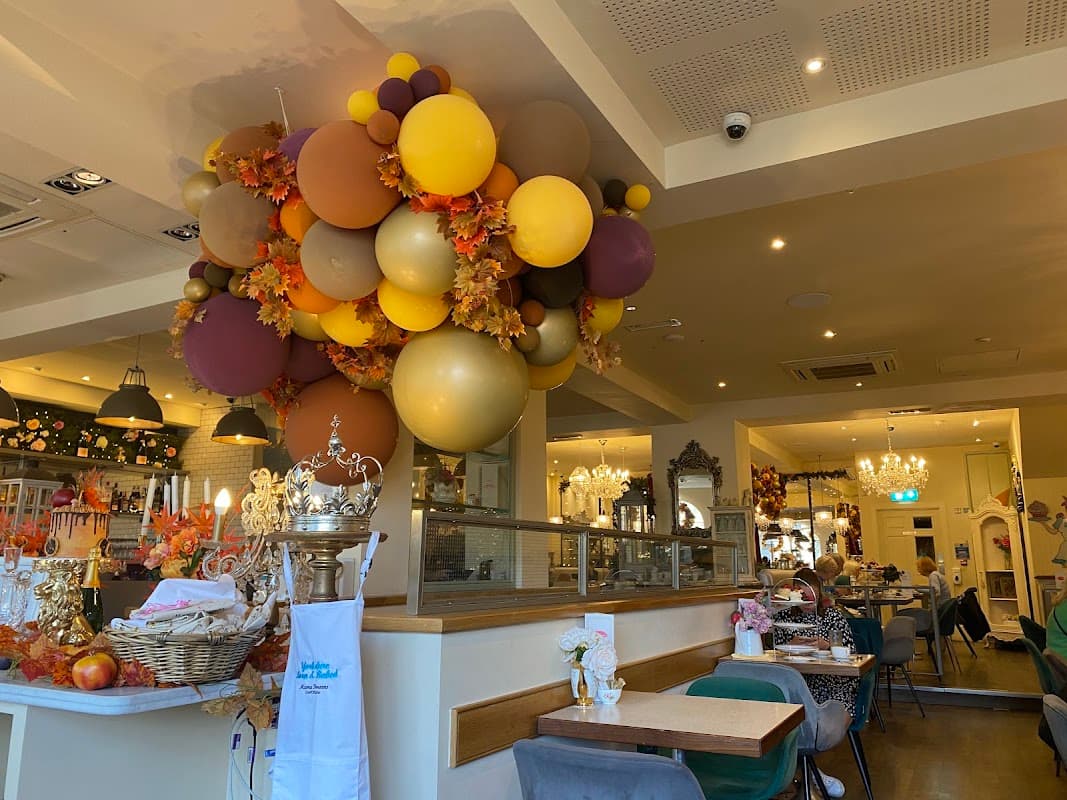 Colorful balloon arrangement hangs above a cozy bakery interior with tables, decor, and patrons enjoying their meals.