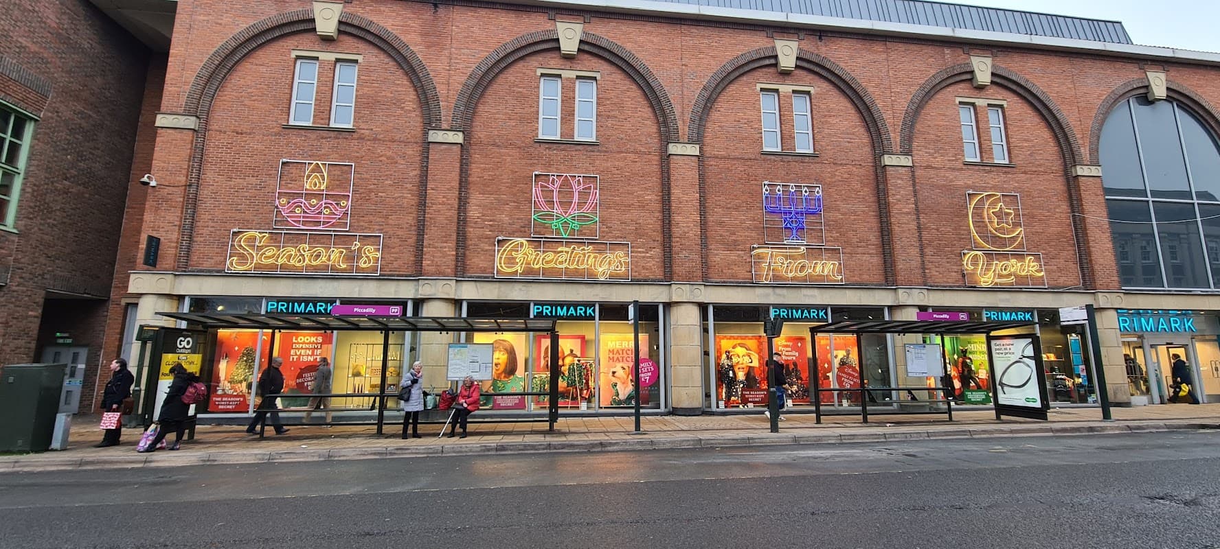 Primark storefront in Harrogate with colorful neon signs and shoppers outside, featuring large windows and brick architecture.
