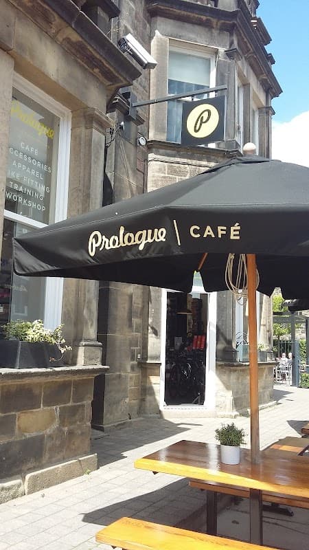 A black umbrella with "Prologue Café" text, outdoor seating, and a stone building with bike accessories displayed.