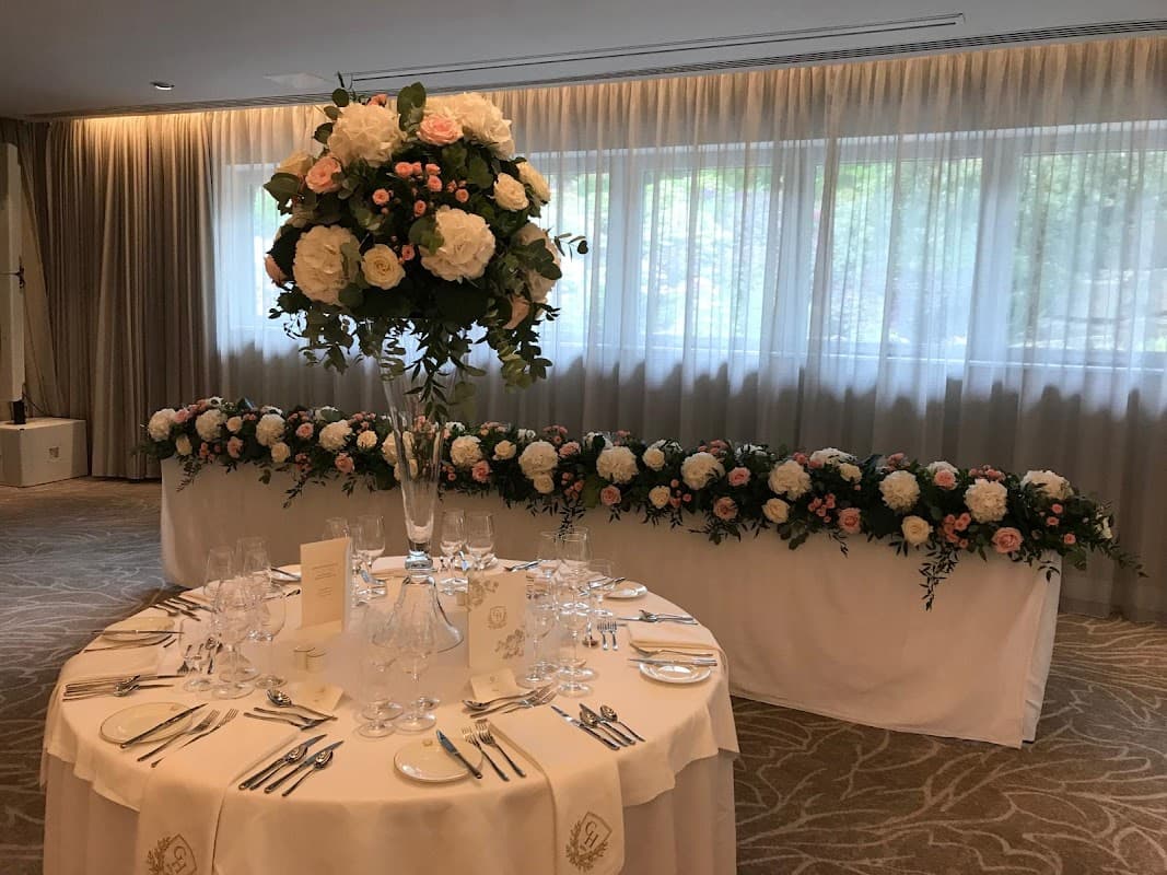 Elegant floral arrangements in tall vases and along a table, set in a softly lit room with sheer curtains.