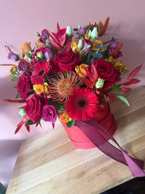 Vibrant floral arrangement in a red bucket, featuring roses, gerberas, and tropical blooms against a pink background.