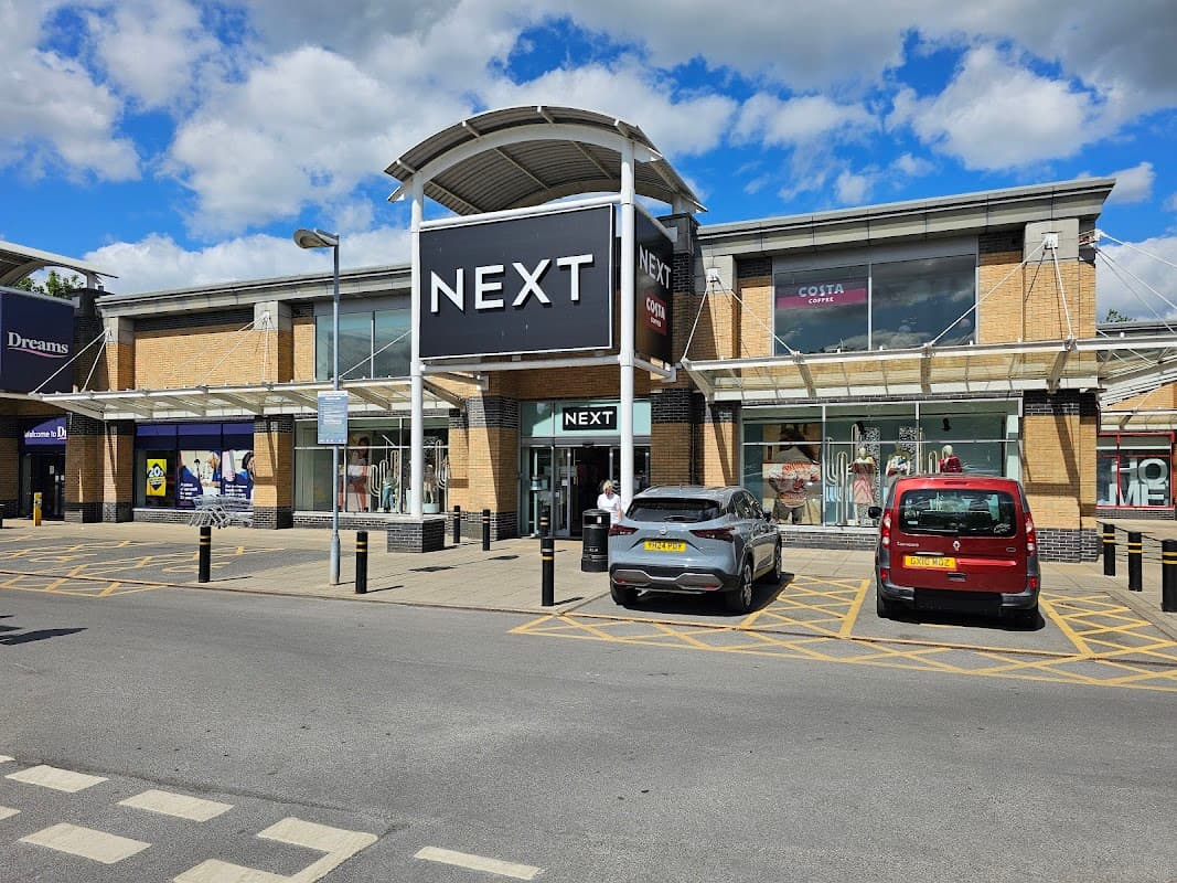 NEXT store at St James Retail Park, with parked cars and a blue sky in the background.