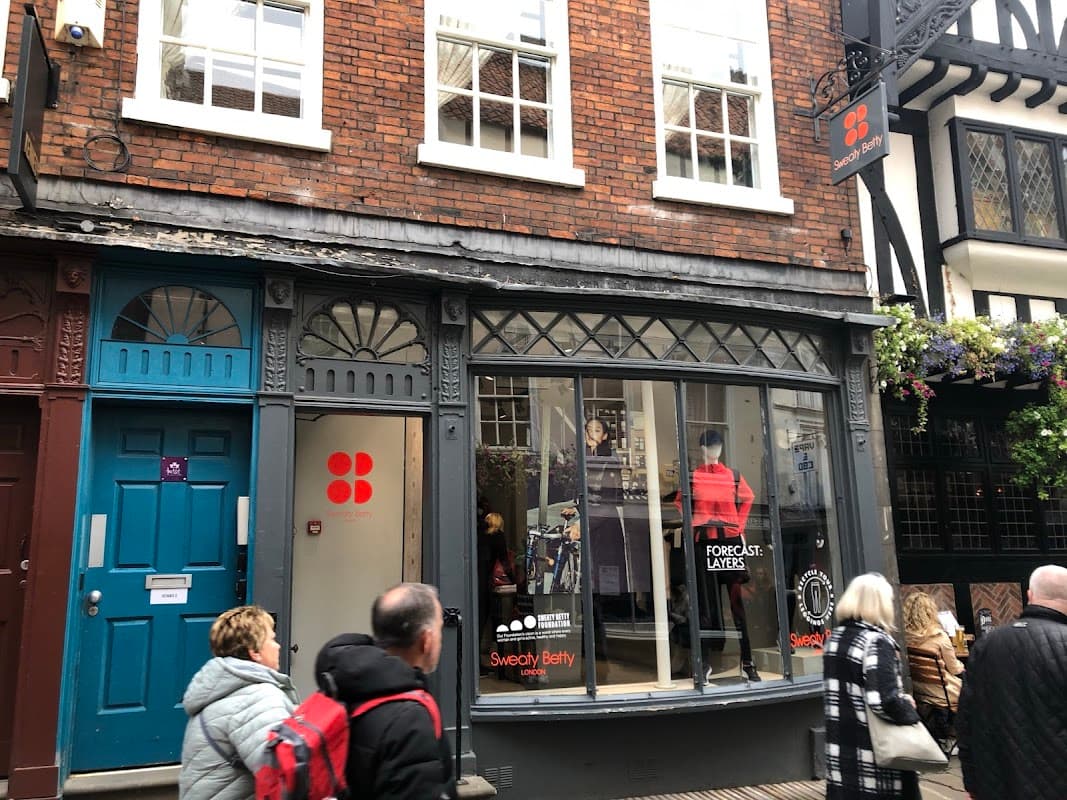 Sweaty Betty clothing store with large front window, red and black signage, and shoppers walking by in a quaint street.